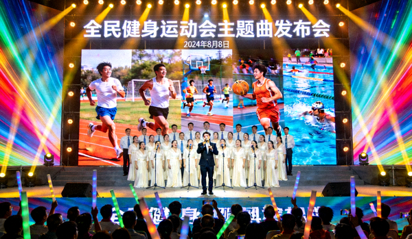 全民健身运动会主题曲发布会现场 - 歌手与合唱团共同演绎，背景大屏幕播放运动画面